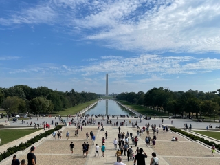 A Large Group Of People Walking Around A Large Courtyard With A Tall Tower In The Background With National Mall In The Background