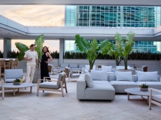 A Man And Woman Standing In A Room With White Couches And A Large Glass Wall With Plants